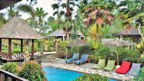 Zona de piscina con sillas de descanso y sombrillas, rodeada de exuberante vegetación tropical.