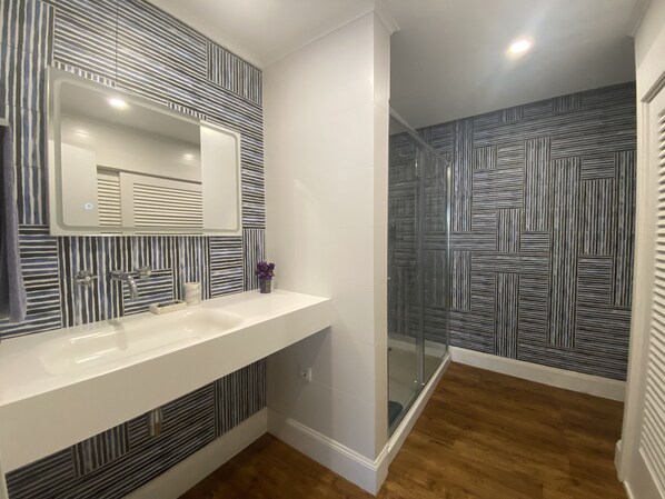 Un baño moderno con tocador blanco, un espejo grande y una ducha con pared decorada.