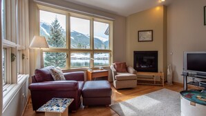 Un salon comprenant un canapé, un pouf et un fauteuil, une cheminée, un téléviseur et une grande fenêtre donnant sur les montagnes.