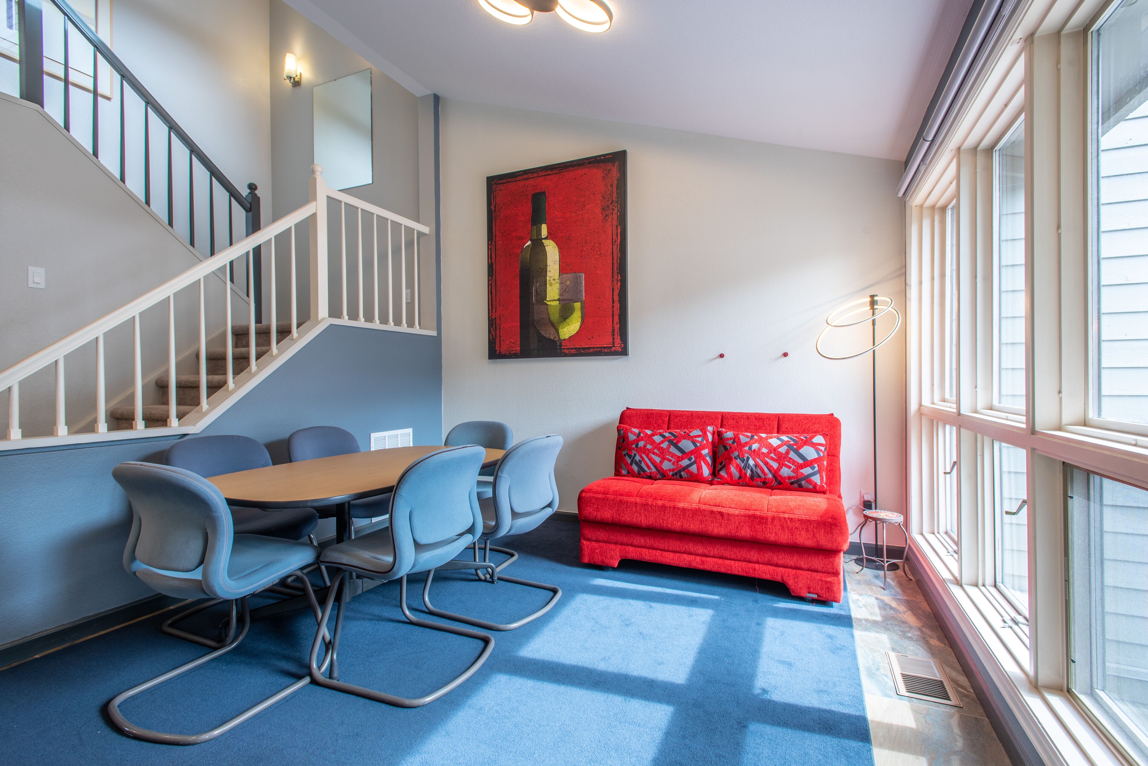 A room with a blue carpet, a red sofa, a dining table with chairs, and a staircase.
