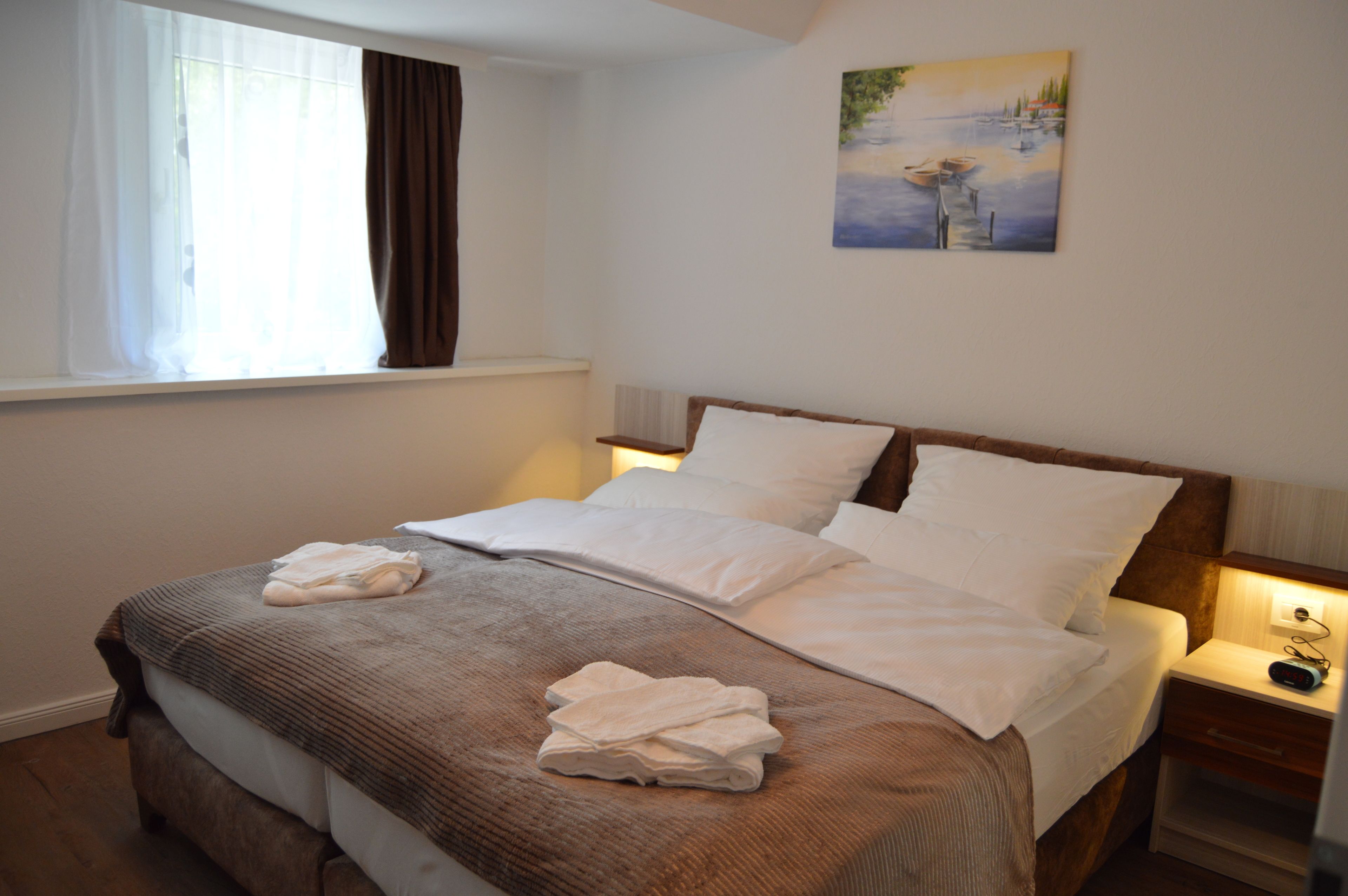 Ein ordentlich bezogenes Bett mit weißer Bettwäsche und einem braunen Kopfteil, ein Nachttisch mit Lampe sowie ein Bild von Booten auf einem See an der Wand.
