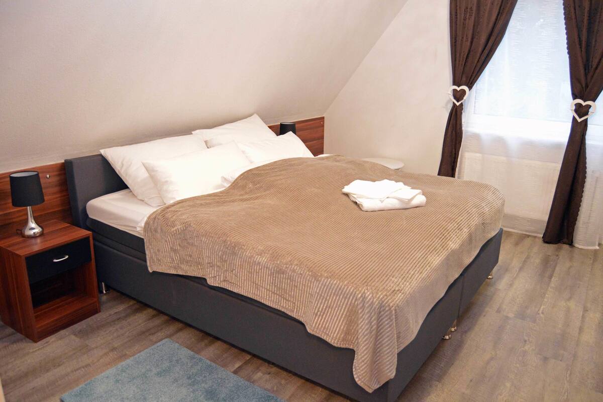 Una cama bien tendida con una cubierta marrón, una mesita de noche con una lámpara y una alfombra azul sobre un suelo de madera.
