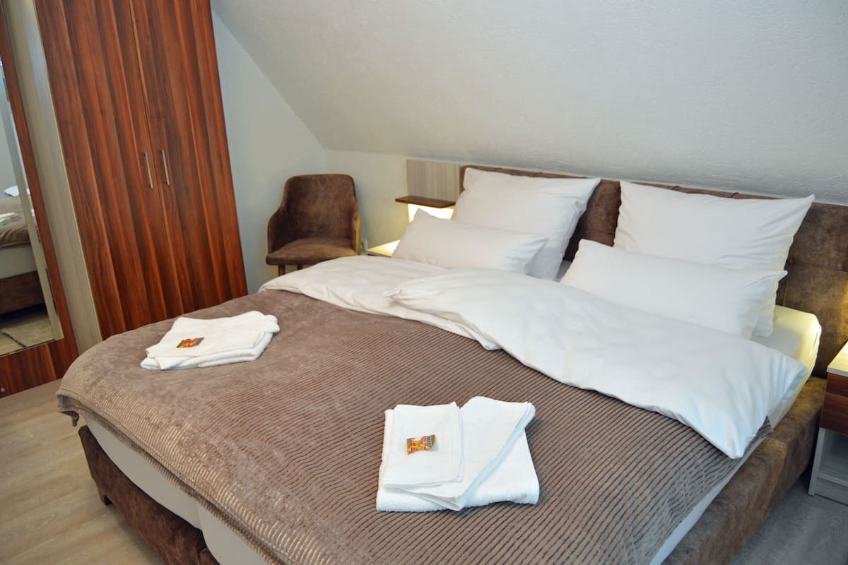 Una cama bien tendida con sábanas blancas y una manta marrón, un armario de madera, un sillón marrón y una mesita de noche con una lámpara.