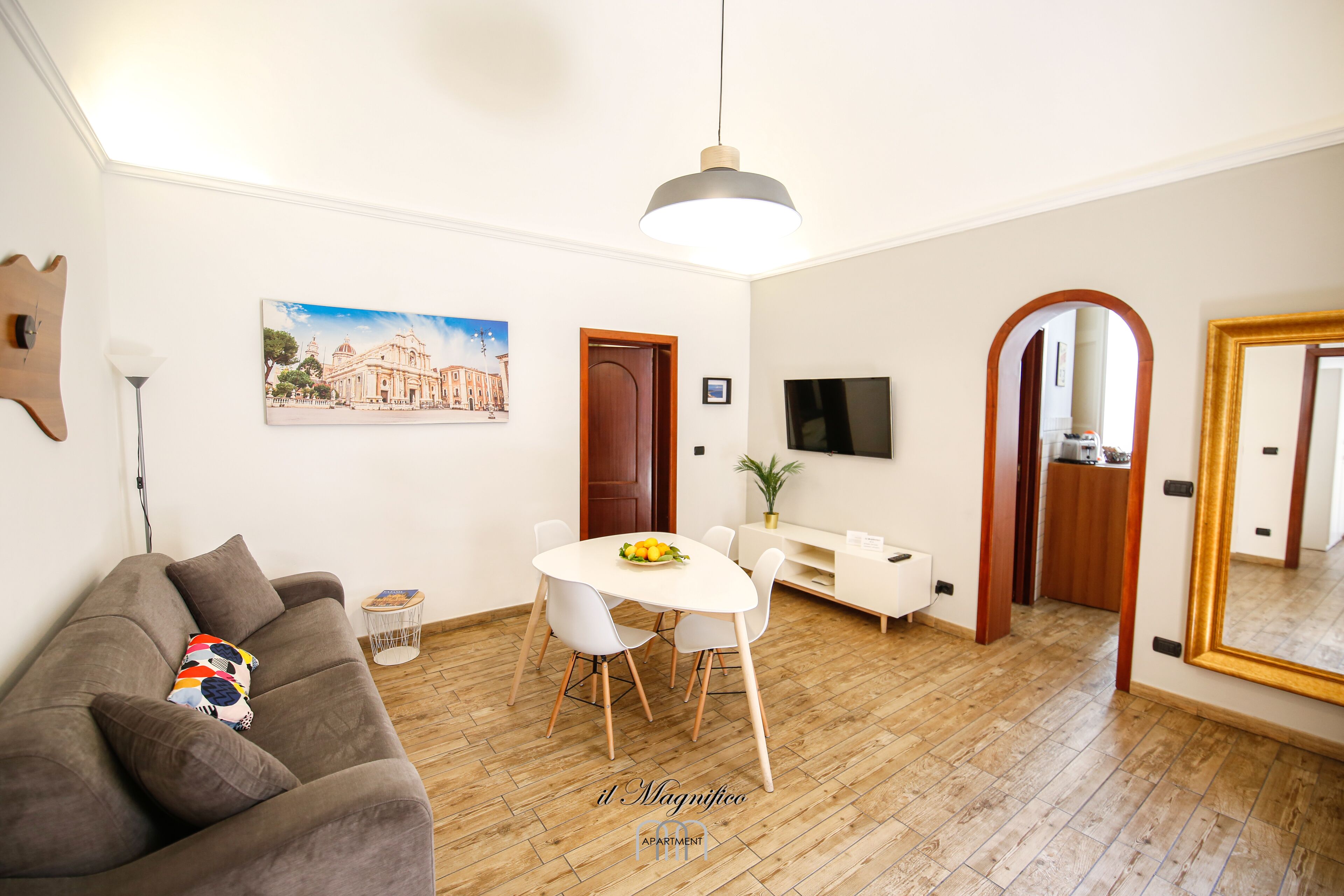Ein modernes Wohnzimmer mit Sofa, Esstisch und Stühlen. An der Wand hängt ein Gemälde, es gibt einen Fernseher und einen großen Spiegel.