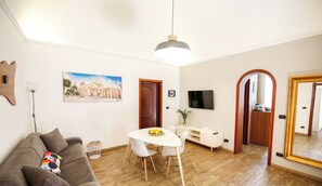 A modern living room with a sofa, dining table, and chairs. There is a painting on the wall, a TV, and a large mirror.