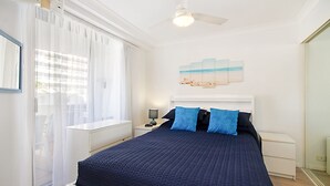 Una camera da letto con un letto, un comodino, una lampada, una ventola a soffitto e una finestra con tende leggere.