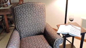 A patterned armchair, a table with a book and a glass of wine, and a floor lamp.