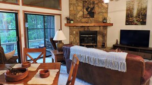 A cozy living room with a stone fireplace, a television, and a large window overlooking a deck.