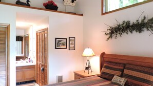 A bedroom with a bed, a nightstand, a lamp, a window with a view of trees, a door, and a mirror.