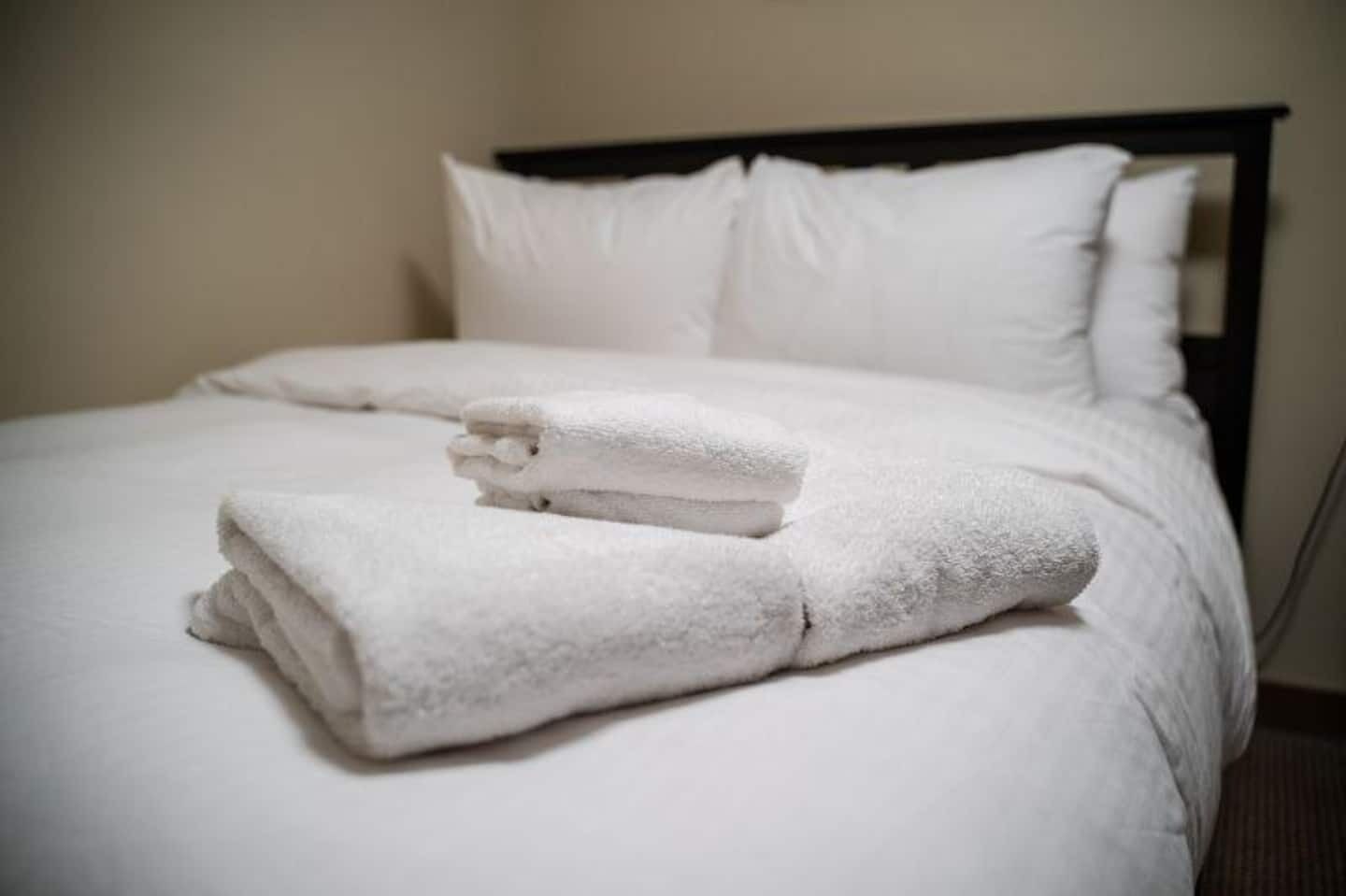 Een netjes opgemaakt bed met witte lakens en kussens, een gevouwen witte handdoek op het bed, en een donker hoofdbord.