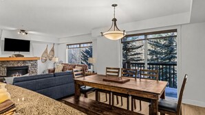 Una sala moderna con chimenea, comedor con mesa y sillas de madera, ventana grande con vista a árboles y televisor de pantalla plana sobre la chimenea.