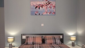 Un lit bien fait, avec une couette à carreaux, deux lampes de chevet et un tableau représentant des flamants roses accroché au mur.