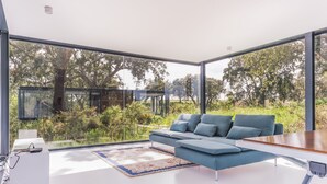 A modern living room with a blue sofa, a wooden coffee table, and a large window offering a view of greenery and trees. - Bungalow 2 - Montemor (Foros de Vale Figueira)