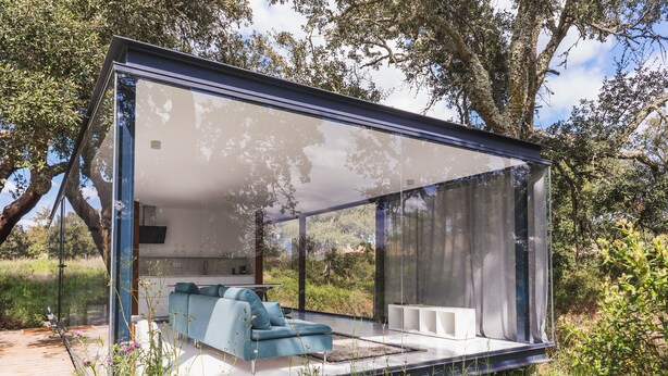 A modern, glass-enclosed structure with a sofa and a small table, surrounded by natural vegetation. - Bungalow 2 - Montemor (Foros de Vale Figueira)