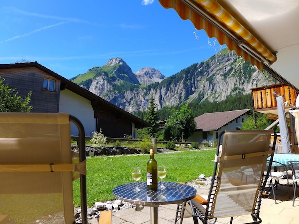 A patio with a table, chairs, and a bottle of wine, overlooking a mountainous landscape.