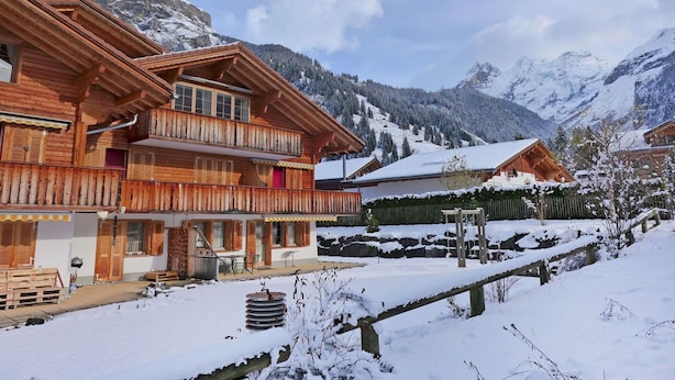 A wooden chalet with a balcony, a covered outdoor seating area, and a view of snow-covered mountains.