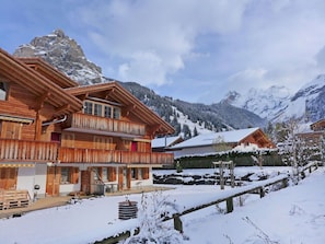 A wooden chalet with a balcony, a covered outdoor seating area, and a view of snow-covered mountains.