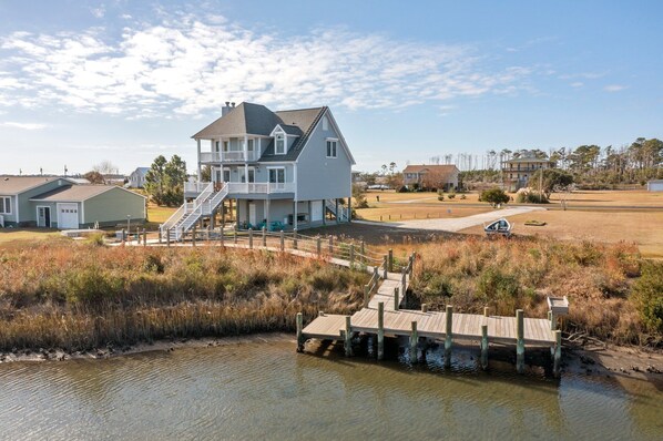 A two-story house with a dock extending into a body of water, surrounded by grassy areas and other houses.