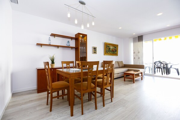 A modern living room with a dining area, featuring a wooden dining table, chairs, a sofa, and a coffee table. There are shelves with decorative items and a painting on the wall.