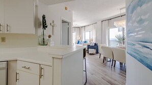 A spacious living area with a dining table, a kitchen area with white cabinetry, and a large painting on the wall.