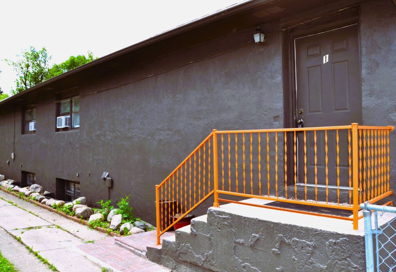 A building with a dark exterior, a single door, and a small set of steps leading to a porch with an orange railing.