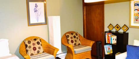 A hotel room with two wicker chairs with patterned cushions, a bed, a bedside table with a lamp, a bookshelf with books, and a framed picture on the wall.