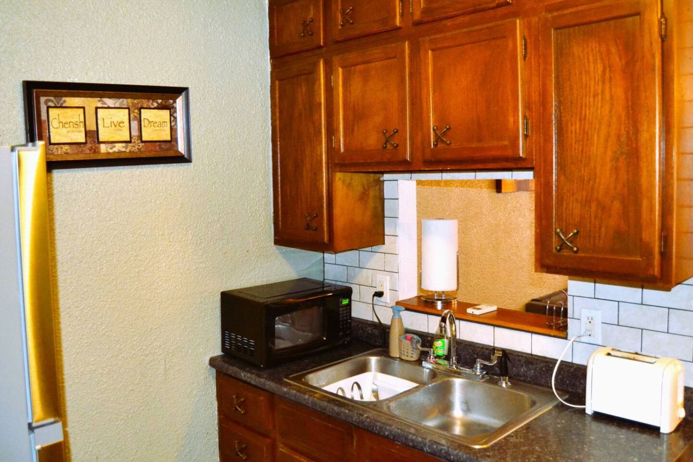 A kitchen with wooden cabinets, a microwave, a sink, a toaster, and a framed inspirational quote on the wall.