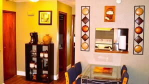 Un comedor con una mesa y sillas de vidrio, una zona de cocina con estufa y armarios, y una estantería con objetos decorativos.
