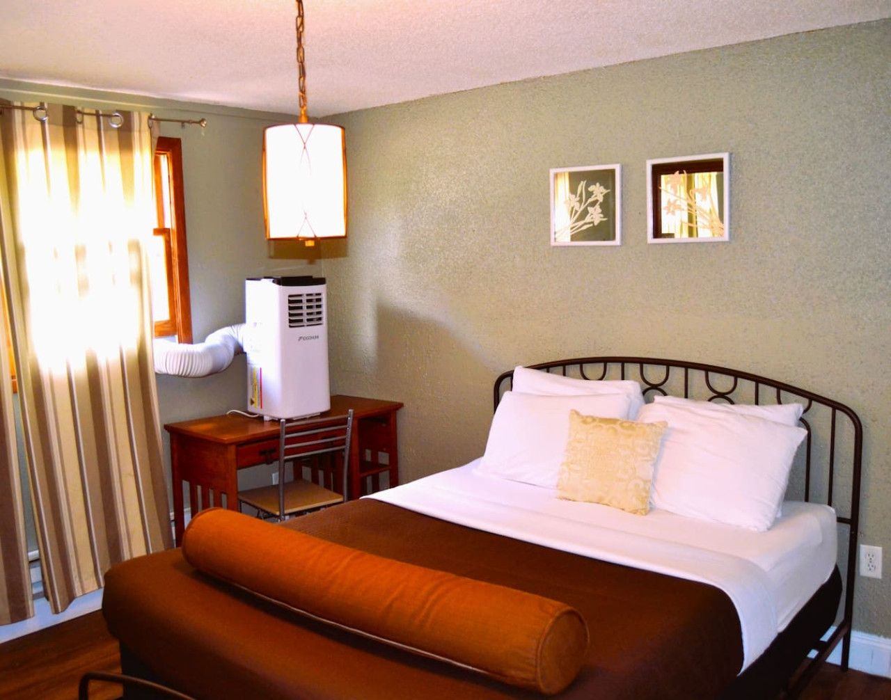 A hotel room with a bed, a desk, a chair, a heater, and a window with curtains.