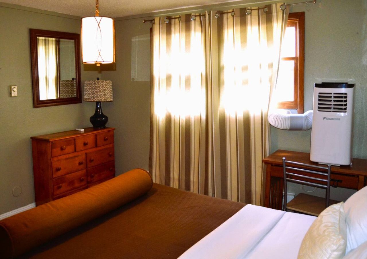 Una habitación de hotel con una cama, un aparador, un escritorio con silla, un calentador portátil y una ventana con cortinas.