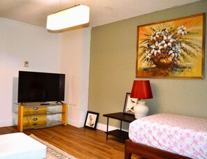 Une chambre avec un téléviseur posé sur un meuble en verre, un lit recouvert d’une couvre-lit à motifs floraux, un tableau accroché au mur et une lampe rouge sur une table de chevet.