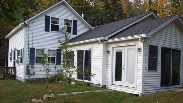 A two-story white house with a dark roof, a front porch, and a small tree in the yard. - Light Oak Leaf Cottage (Bar Harbor)