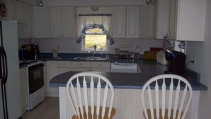 A kitchen with white cabinets, a window with a blue and white valance, a sink, and two wooden chairs with white backs. - Light Oak Leaf Cottage (Bar Harbor)