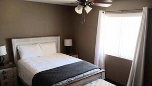 Un dormitorio con cama, mesita de noche, lámpara, ventana con cortinas y ventilador de techo.