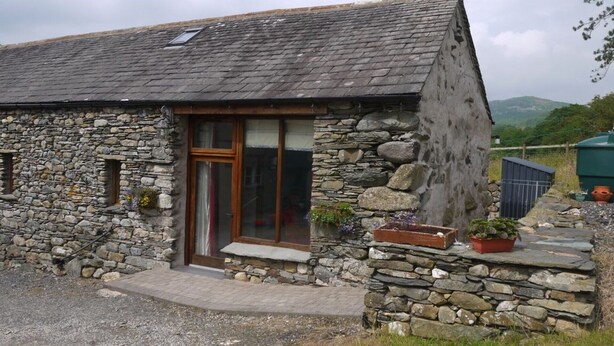 A stone cottage with a sloped roof, a wooden door, and a small porch area with potted plants.