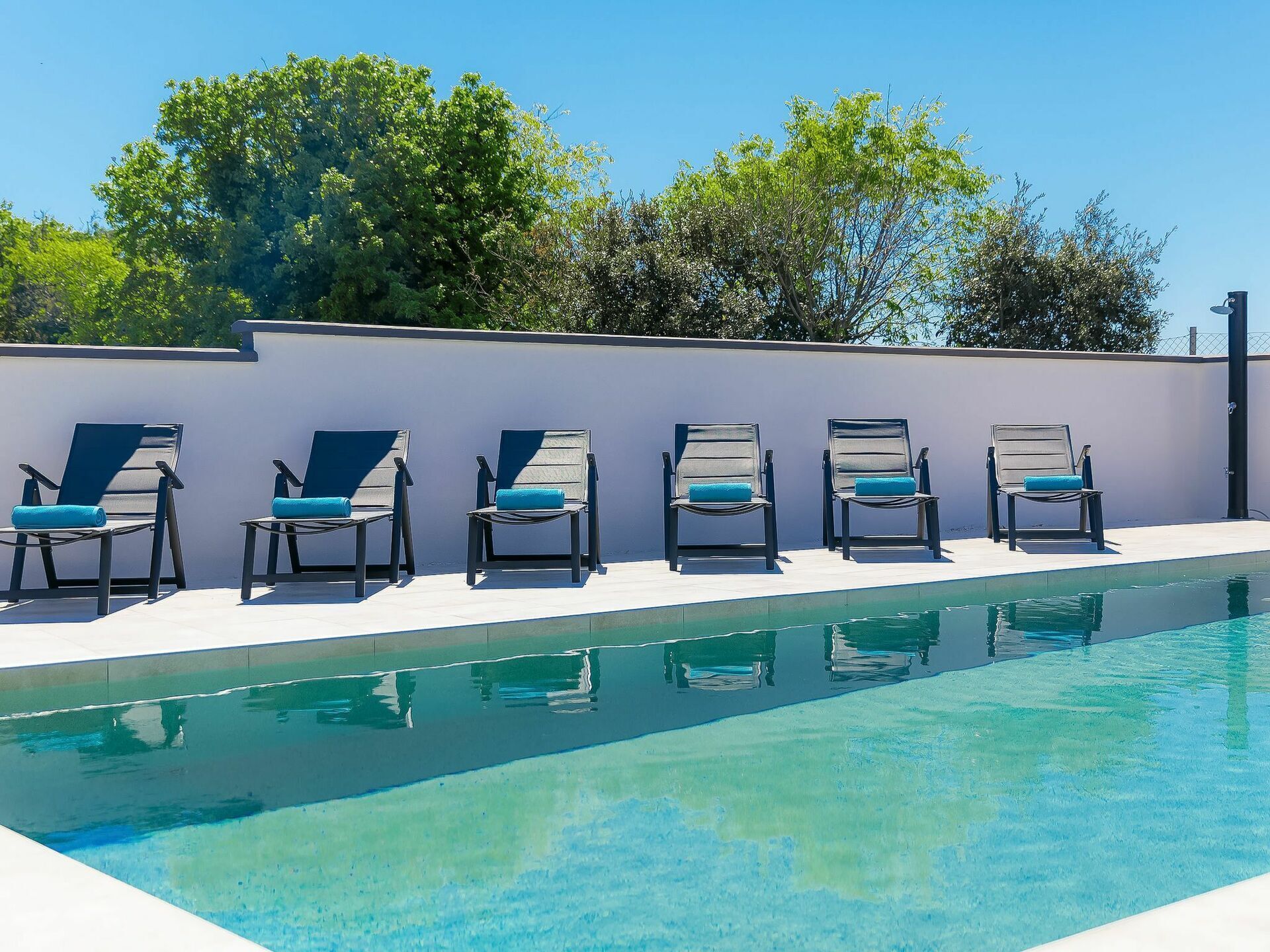 Un área junto a la piscina con seis tumbonas alineadas contra una pared, una piscina azul cristalina y árboles al fondo.