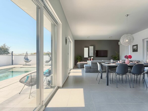 A modern dining area with a pool view, a flat-screen TV, and a large chandelier.