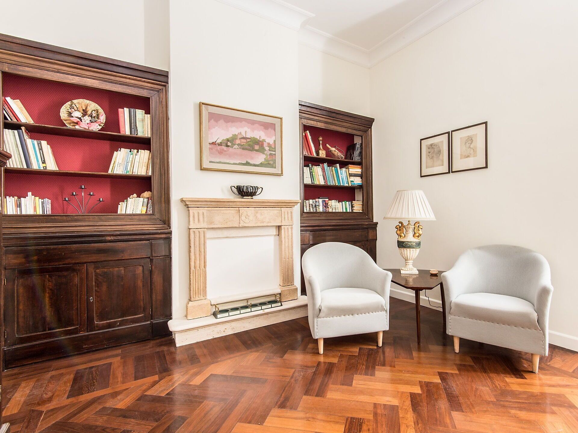 A room with two armchairs, a fireplace, bookshelves, and framed artwork.