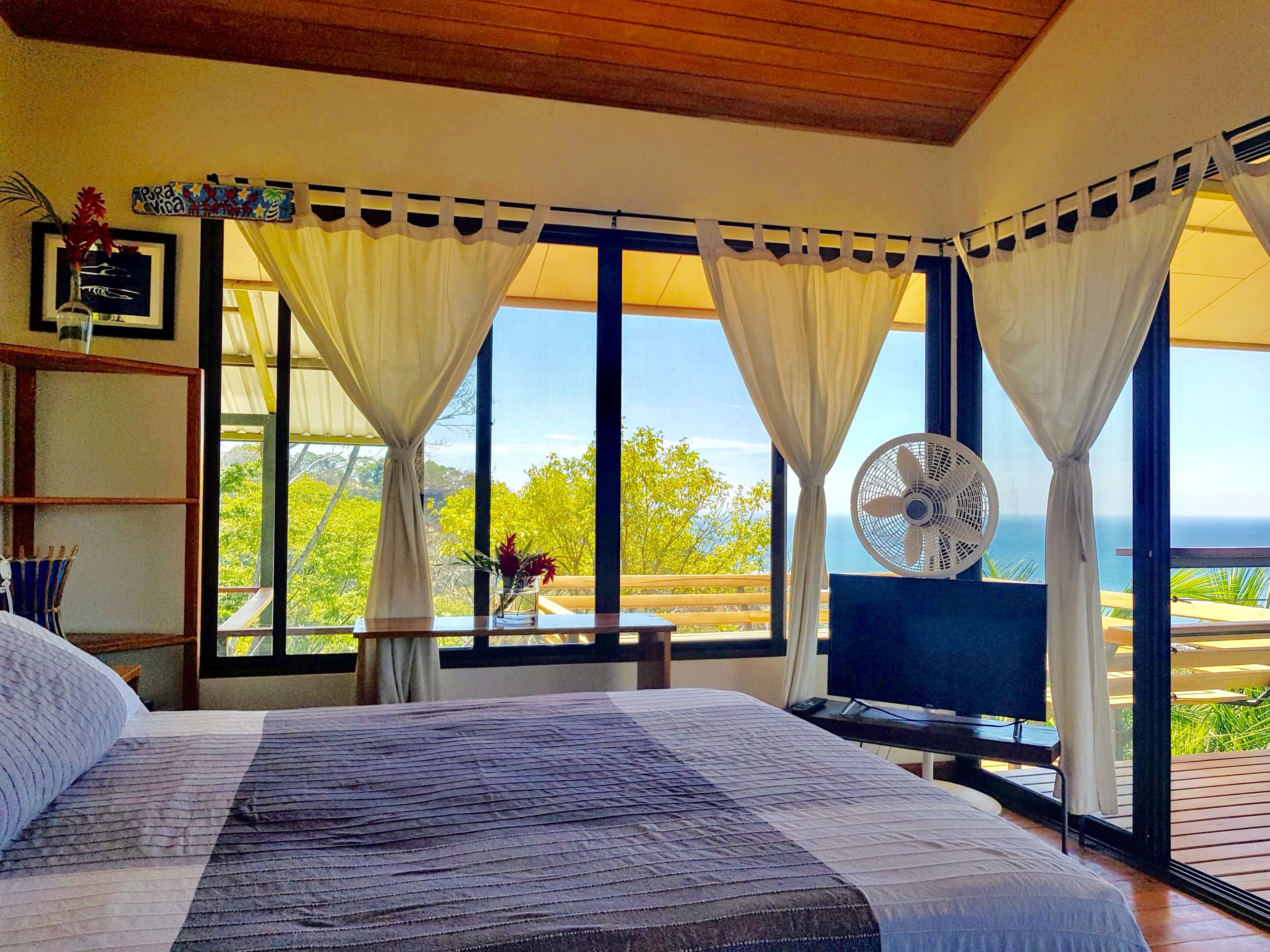 A bedroom with a bed, a desk, a chair, a fan, and a view of the ocean.