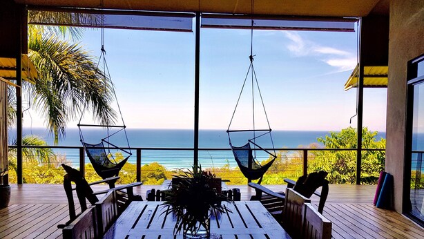 Eine Holzterrasse mit Tisch und Stühlen, zwei Hängematten und Blick auf das Meer.