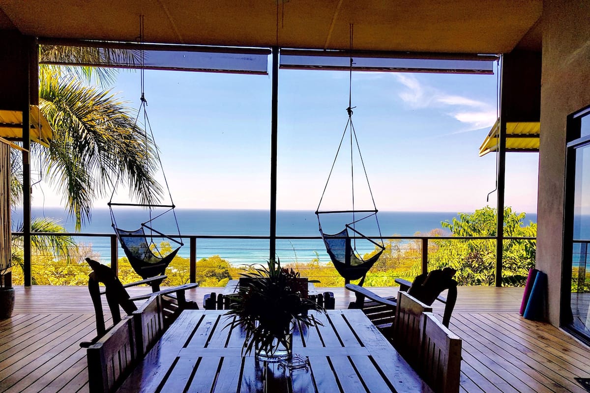 Eine Holzterrasse mit Tisch und Stühlen, zwei Hängematten und Blick auf das Meer.