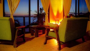 Ein gemütlicher Sitzbereich mit Blick auf das Meer im Dämmerlicht, mit Holzmöbeln und einem Tisch mit Lampe.