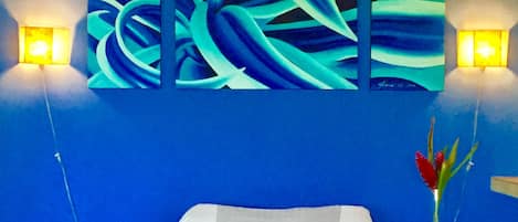 Una habitación con cama, dos lámparas de noche, un cuadro en la pared y una mesita con un jarrón.