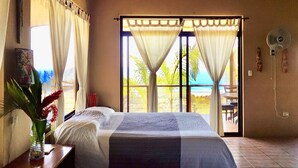 Une chambre à coucher avec un lit, des rideaux, une fenêtre donnant sur la mer, une table de chevet avec un vase et un ventilateur fixé au mur.