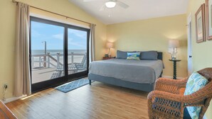 A bedroom with a large bed, a wicker chair, a nightstand, and a view of the beach through a sliding door. - Welcome to Villa Rosa! A 4 Bedroom 3 Bath Oceanfront Getaway (St. Augustine)