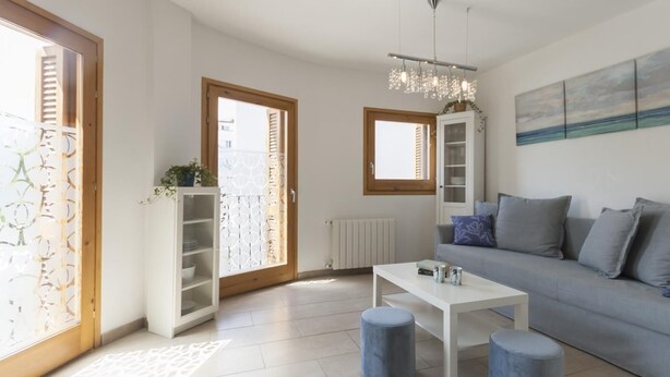 Ein modernes Wohnzimmer mit einem grauen Sofa, einem weißen Couchtisch und zwei blauen Hocker. Eine Holztür mit Milchglasfenster, ein weißes Schränkchen und ein Heizkörper an der Wand sind ebenfalls zu sehen.