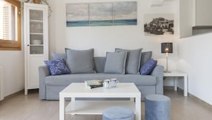 Ein modernes Wohnzimmer mit einem grauen Sofa, einem weißen Couchtisch und einem blauen Hocker. Über dem Sofa hängt ein Gemälde mit einer Strandansicht und auf einem Beistelltisch steht eine Lampe.