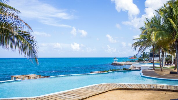 A resort pool with a wooden deck, palm trees, and a view of the ocean. - Whispering Seas (Tower Isle)