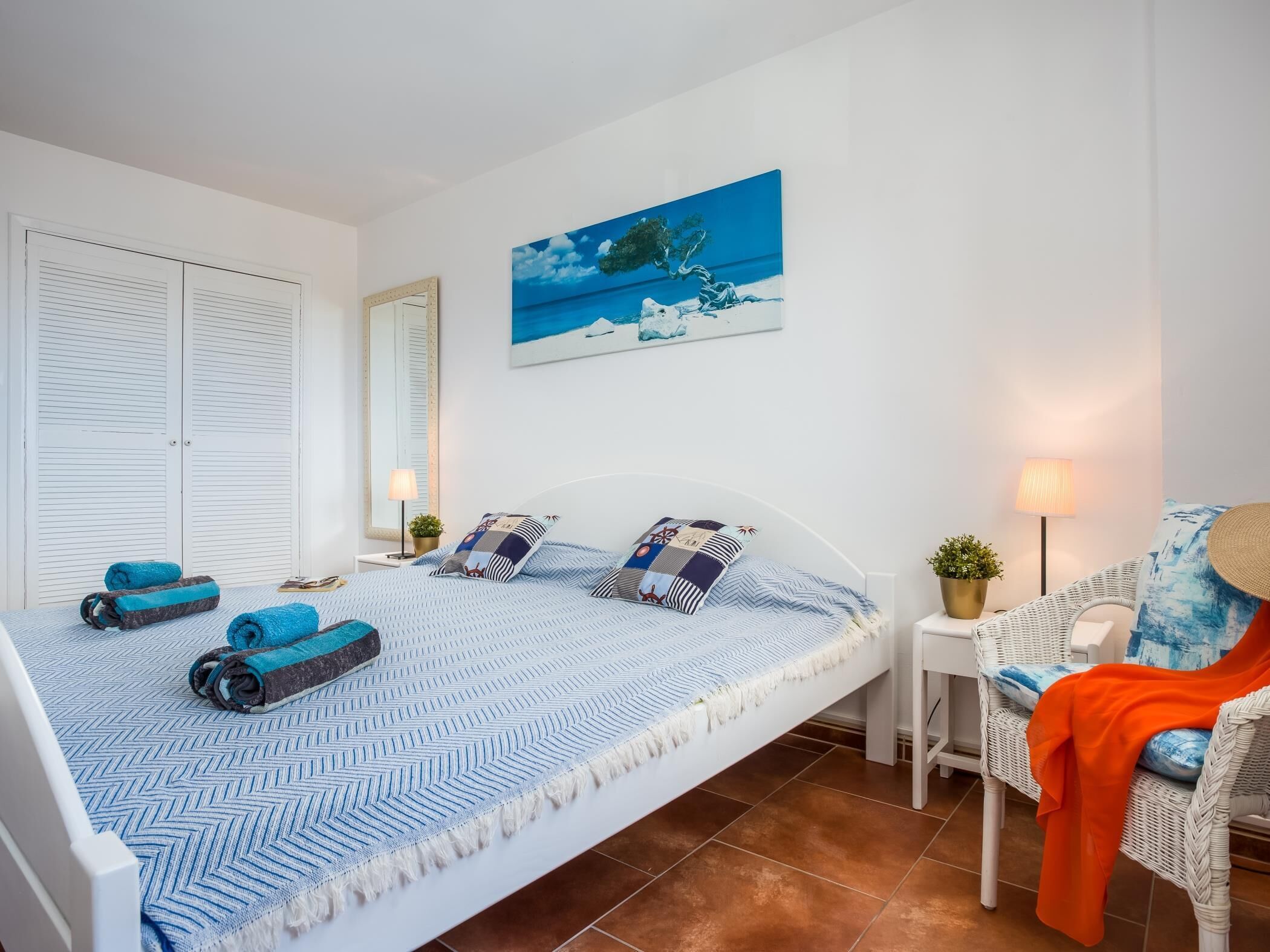 Un dormitorio con cama, una silla, una mesa, una lámpara y un cuadro en la pared.