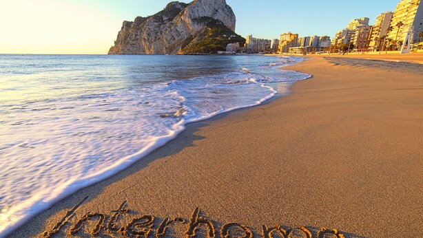 A beach with the word "interhome" written in the sand, a rocky cliff, and buildings in the background. - Cosy apartment for 4 people with pool, A/C, WIFI, TV and terrace (Benissa)